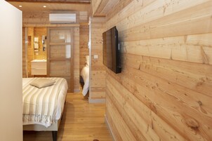 Quarto com painéis de madeira, uma cama, televisão na parede e banheiro visível através de uma porta aberta.