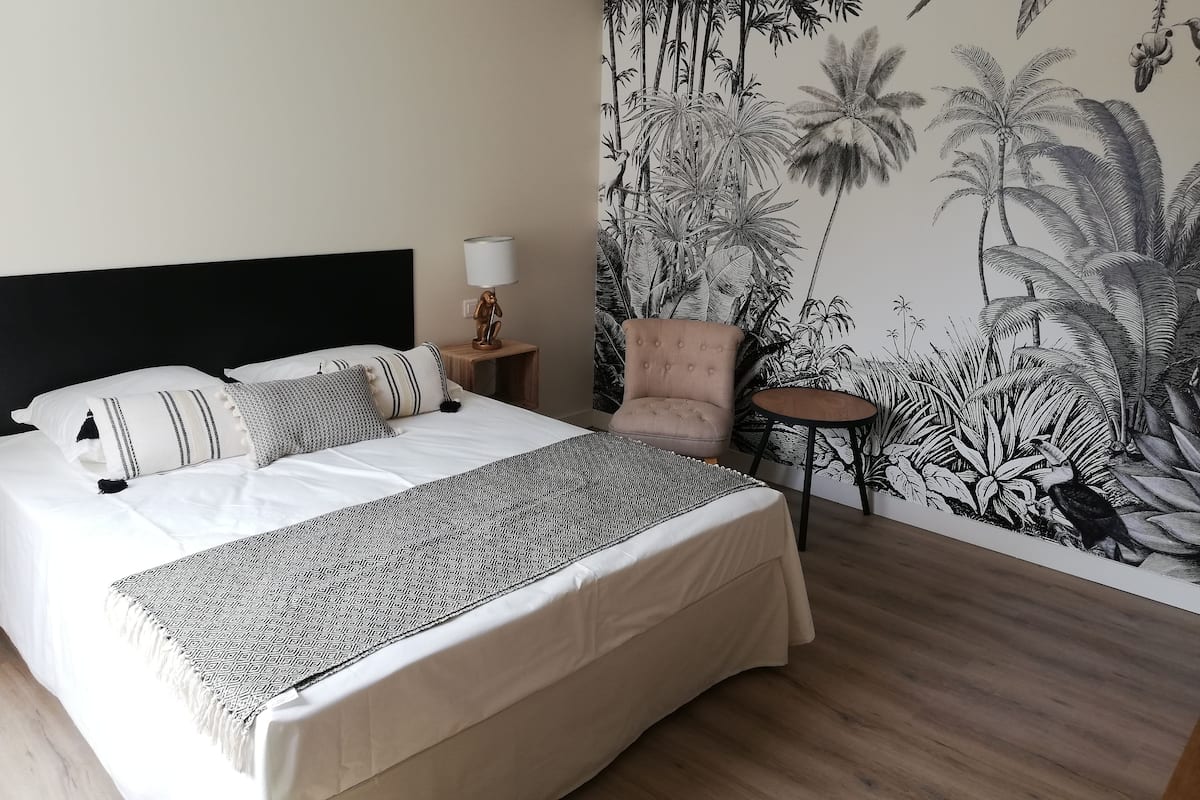Ein Schlafzimmer mit einem großen Bett, einem Nachttisch, einem Sessel, einem kleinen Tisch und einer Wandmalerei mit tropischen Pflanzen.