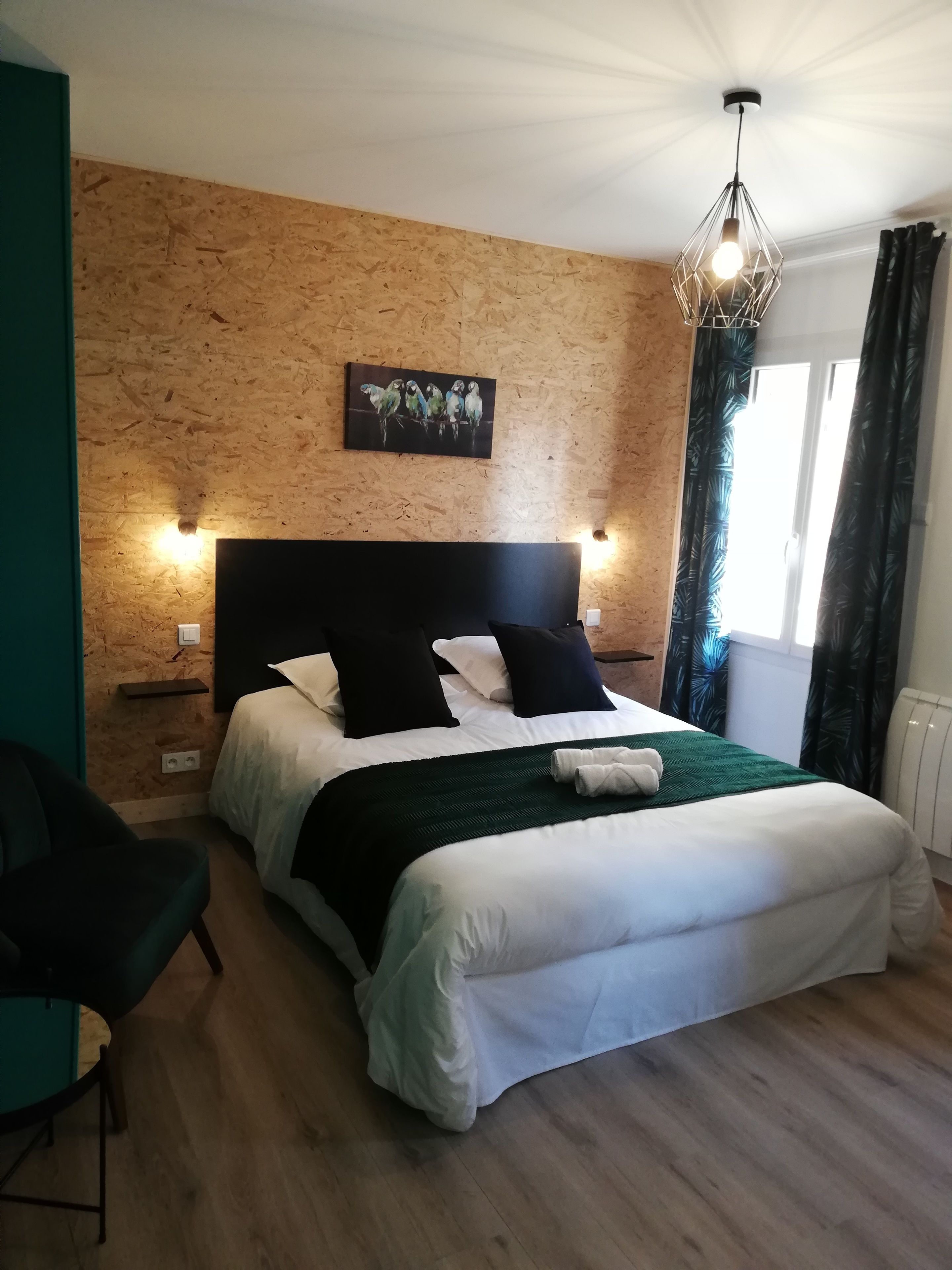 Ein Schlafzimmer mit einer Holzwand, einem Bett mit weißer und schwarzer Bettwäsche, einer grünen Tagesdecke, einem Kopfteil mit Wandleuchten, einem Papageienbild und einem Fenster mit Vorhängen.