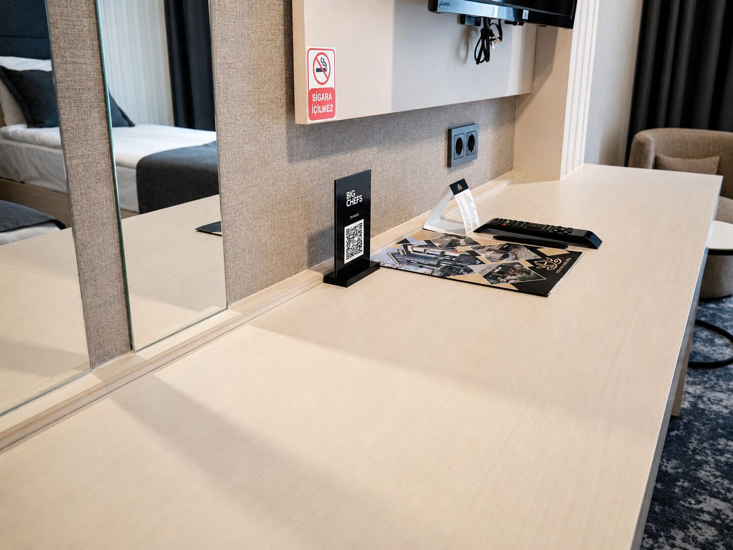 A hotel room with a flat-screen TV mounted on the wall, a desk with a remote control, a QR code sign, and a no-smoking sign.
