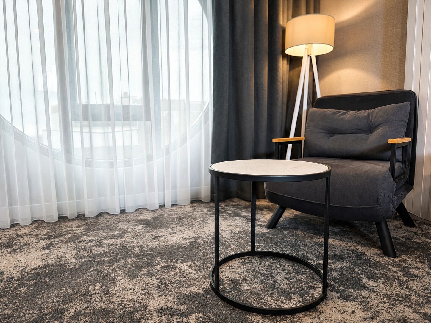 Una habitación con una mesa redonda, un sillón oscuro, una lámpara de pie y cortinas translúcidas.