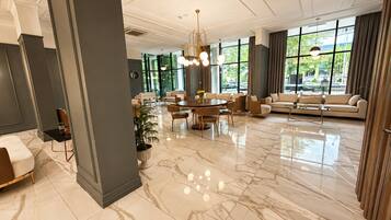 Eine großzügige Hotelhalle mit Marmorboden, einem runden Esstisch, Sofas und großen Fenstern mit Blick ins Freie.