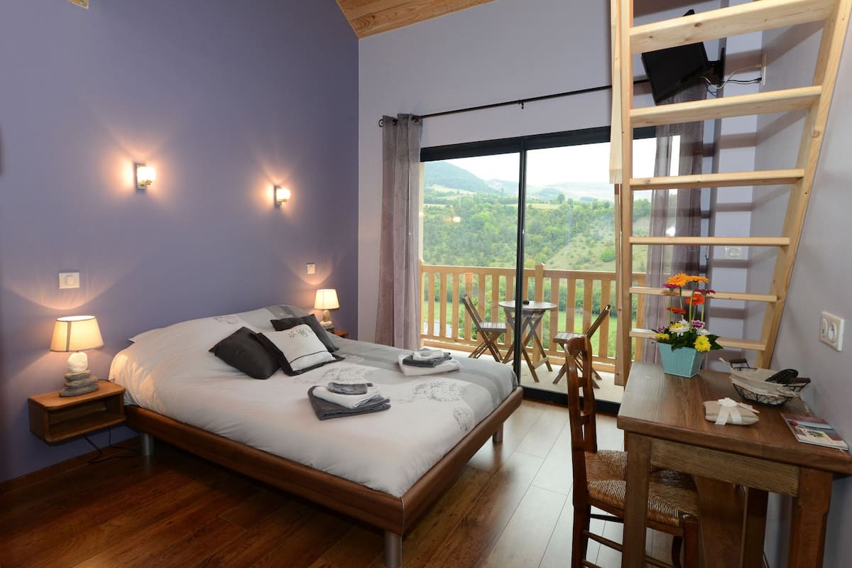 Een slaapkamer met een bed, bedlampen, een houten bureau met een stoel, een raam met uitzicht op groen en een balkon met een leuning.