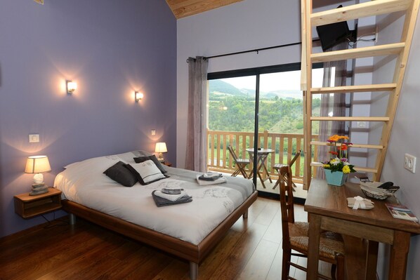 A bedroom with a bed, bedside lamps, a wooden desk with a chair, a window with a view of greenery, and a balcony with a railing. - Lauz'oustal - Maison d'hôtes (Montrodat)
