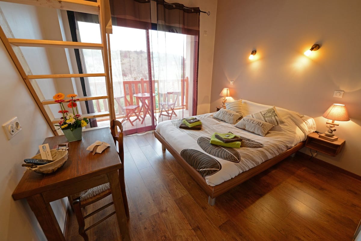 Een slaapkamer met een houten bed, twee bedlampen, een klein tafeltje met een stoel, en een raam met uitzicht op de omgeving.