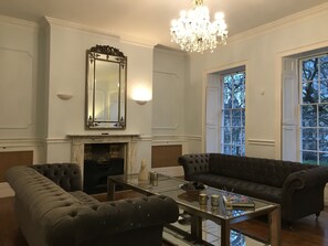 A living room with a fireplace, two dark sofas, a glass coffee table, and a chandelier.