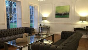 Ein Wohnzimmer mit einer dunkelgrauen Couch, einem Glastisch und Holzboden. Zwei Fenster mit weißen Rahmen, eine Chandelier und ein Gemälde an der Wand sind ebenfalls zu sehen.