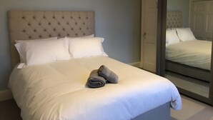 Ein ordentlich bezogenes Bett mit einer gesteppten Kopfteil, weißer Bettwäsche und einem Paar zusammengerollter Handtücher am Fußende.
