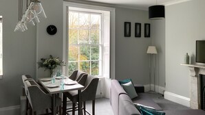 A dining area with a table set for four, a sofa, a fireplace, and a large window with a view of trees. - River View (Bath)