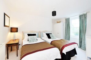Una habitación de hotel con dos camas individuales, una mesita de noche con lámpara, una ventana con cortinas y una puerta.