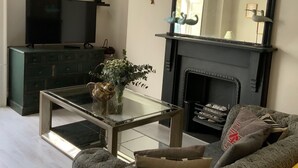 A living room with a sofa, a glass coffee table, a television, a fireplace, and a mirror. - Gilda's Garden (Bath)