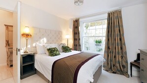 A bedroom with a large bed, two bedside tables with lamps, a window with curtains, and a door leading to another room. - Gilda's Garden (Bath)