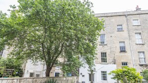 A courtyard with a large tree, outdoor seating, and a building with multiple windows. - Gilda's Garden (Bath)