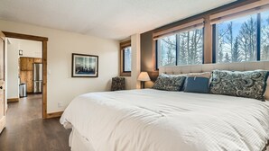 Un dormitorio con una cama grande, una ventana con vistas a árboles, un cuadro enmarcado en la pared y una puerta de madera que da a una cocina.