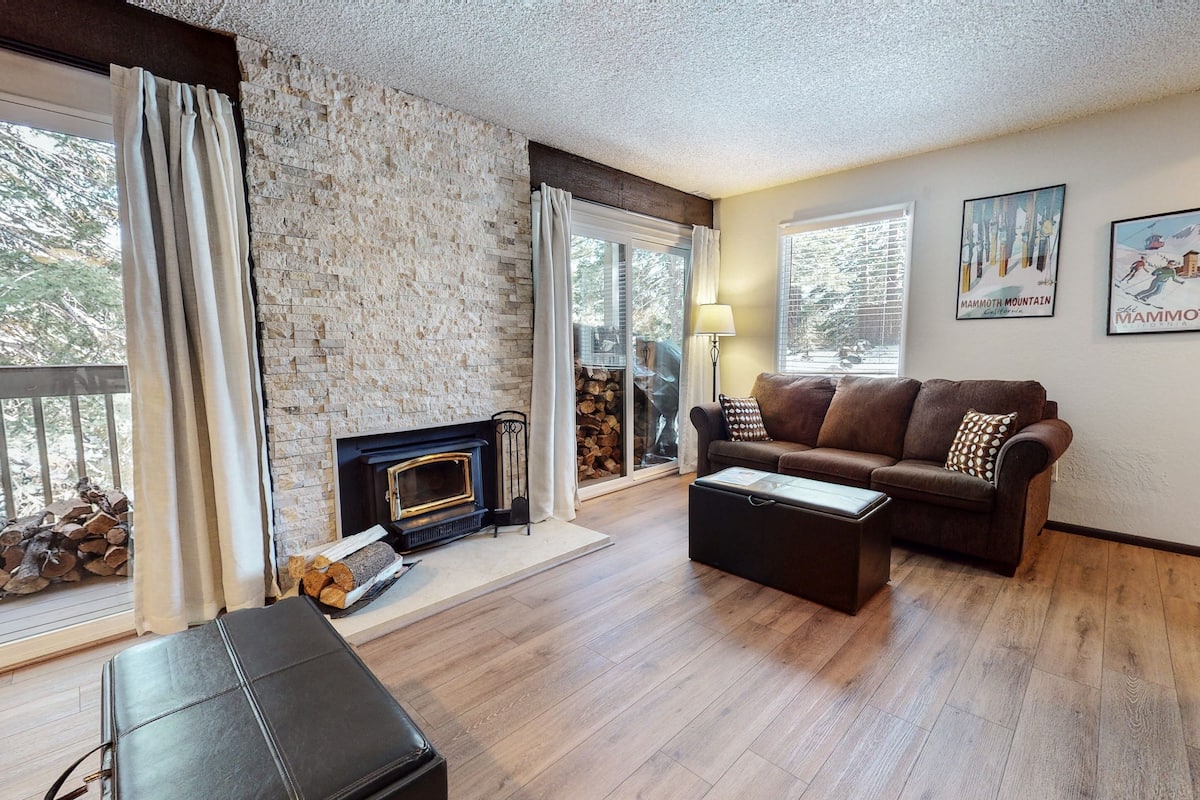 A living room with a fireplace, a sofa, a coffee table, and a bench. There are two windows with curtains, and a lamp is on the table.