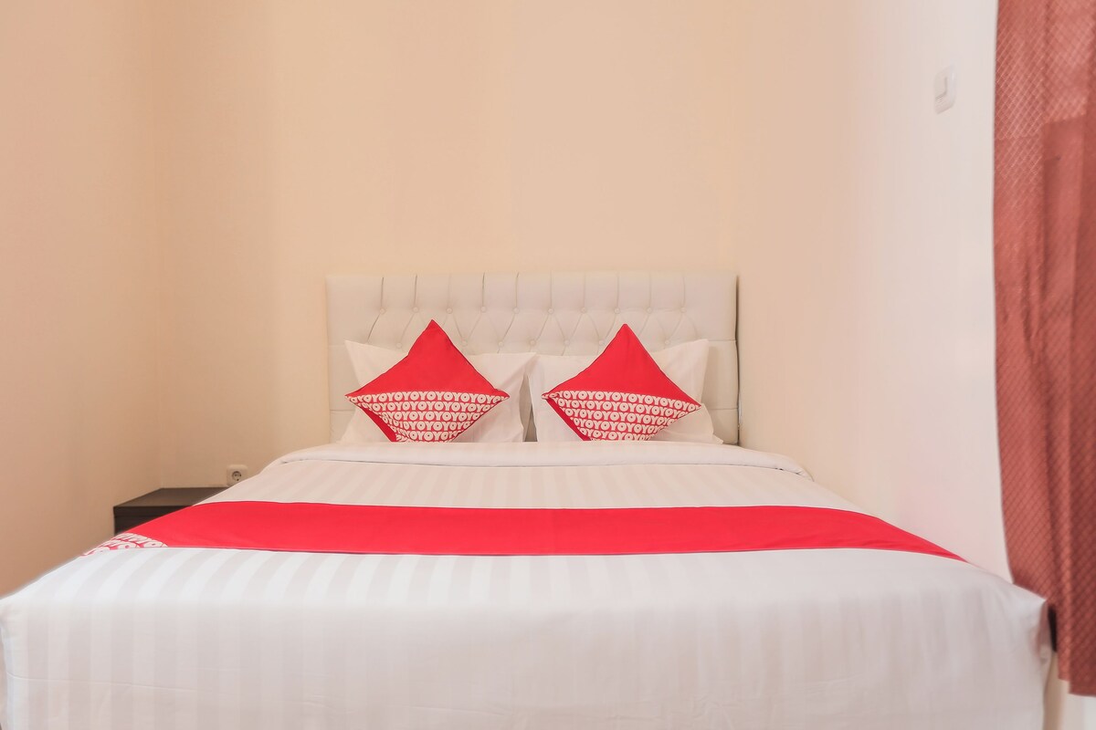 Una cama con cabecera blanca y almohadas rojas, colcha roja y cortina marrón.