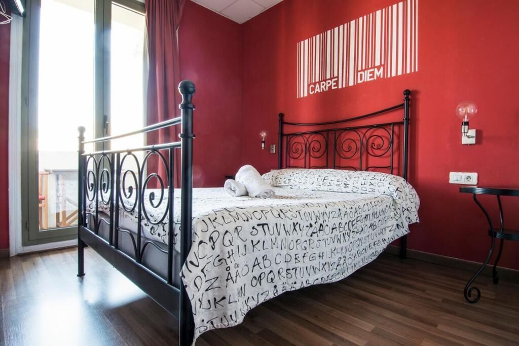 Un dormitorio con estructura de cama metálica, pared roja con diseño de código de barras y una ventana con cortinas.