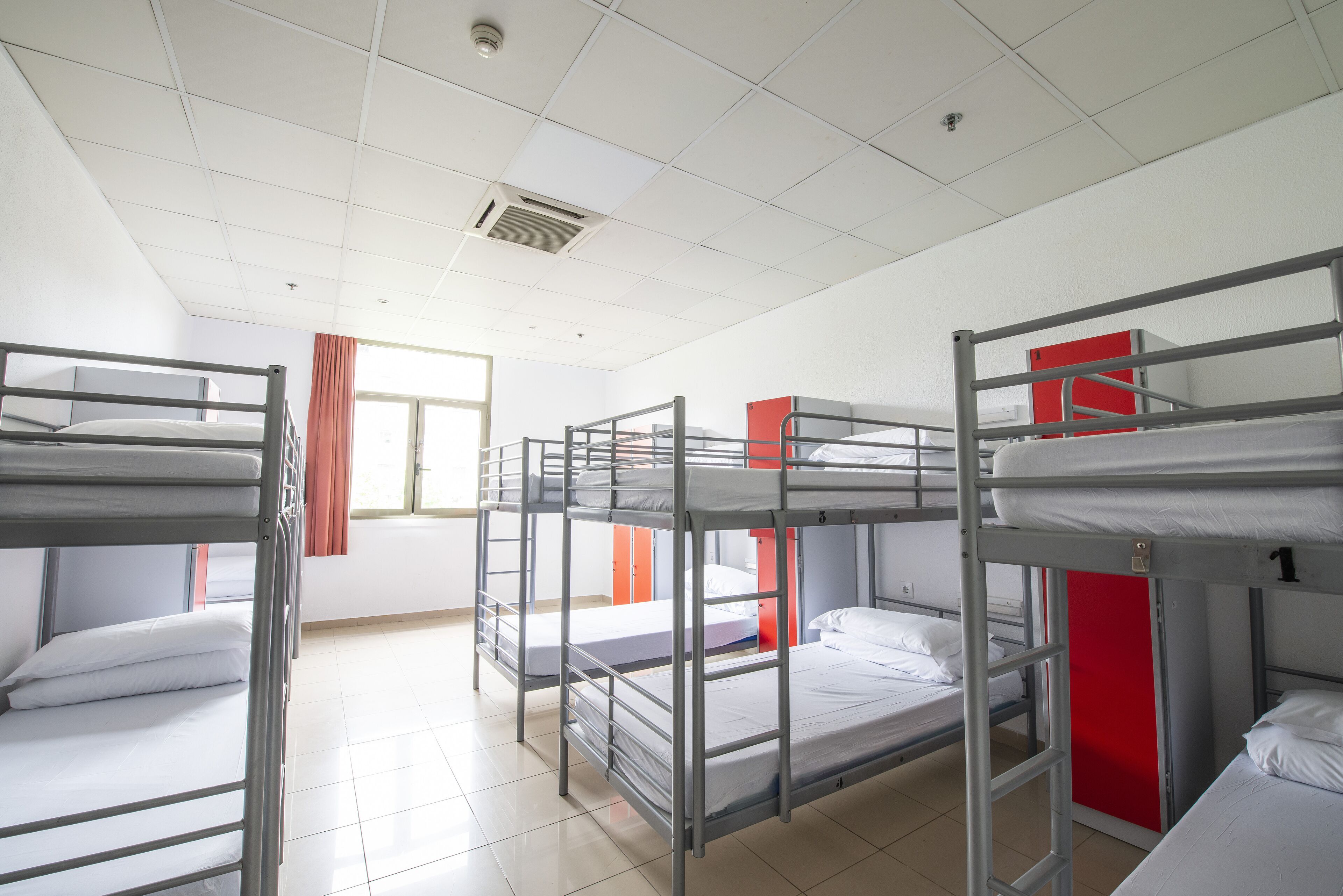 Ein Schlafsaalzimmer mit Etagenbetten, einem Fenster mit Vorhängen und einer roten Tür.