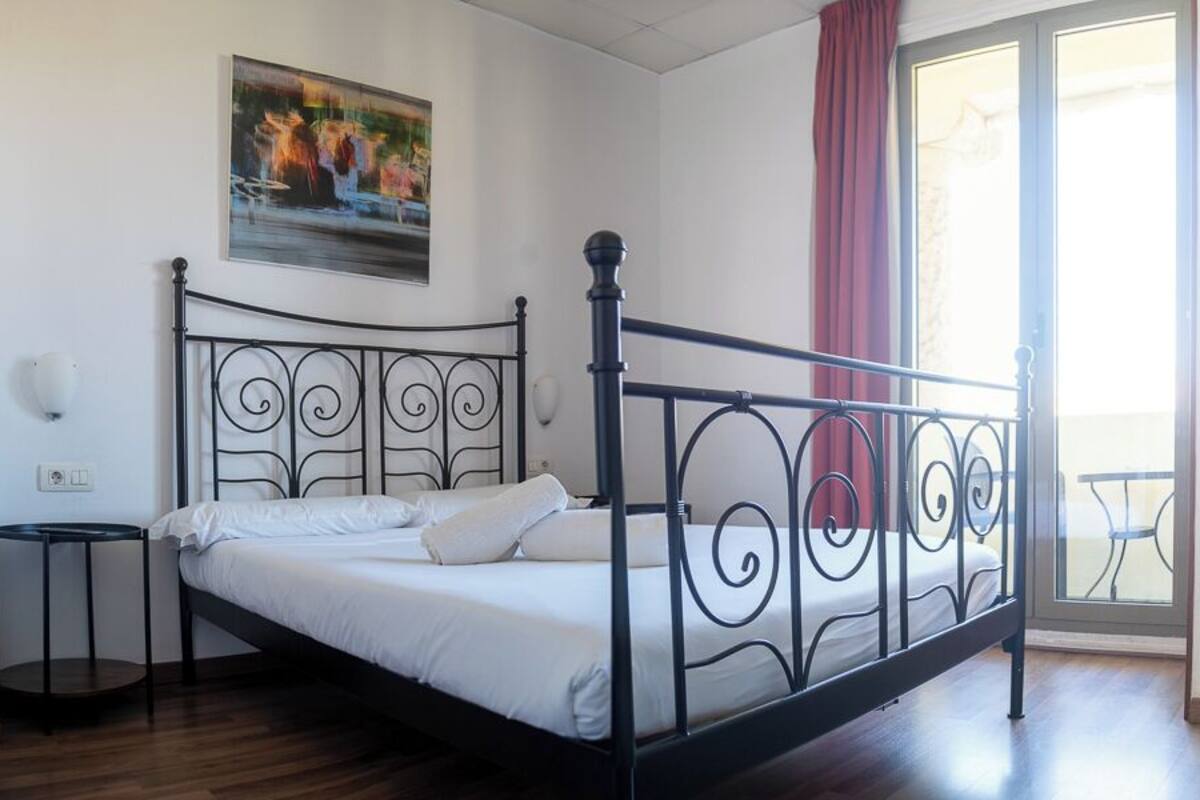 A bedroom with a metal bed frame, white bedding, a small bedside table, a wall-mounted light, and a window with a view.