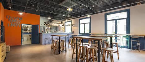 Un grand espace bar avec des tables hautes et des tabourets, une cuisine à l’arrière-plan et une grande fenêtre qui laisse entrer la lumière naturelle.