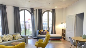 A modern living room with a sofa, armchair, and a bed. There are large windows with curtains, a small table, and a lamp. - Safestay Barcelona Passeig de Gracia (Barcelona)