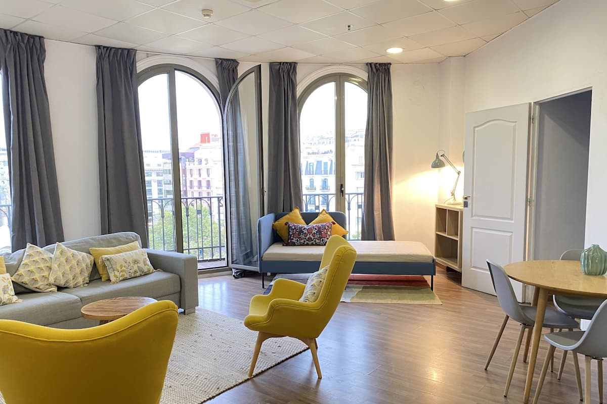 A modern living room with a sofa, armchair, and a bed. There are large windows with curtains, a small table, and a lamp.