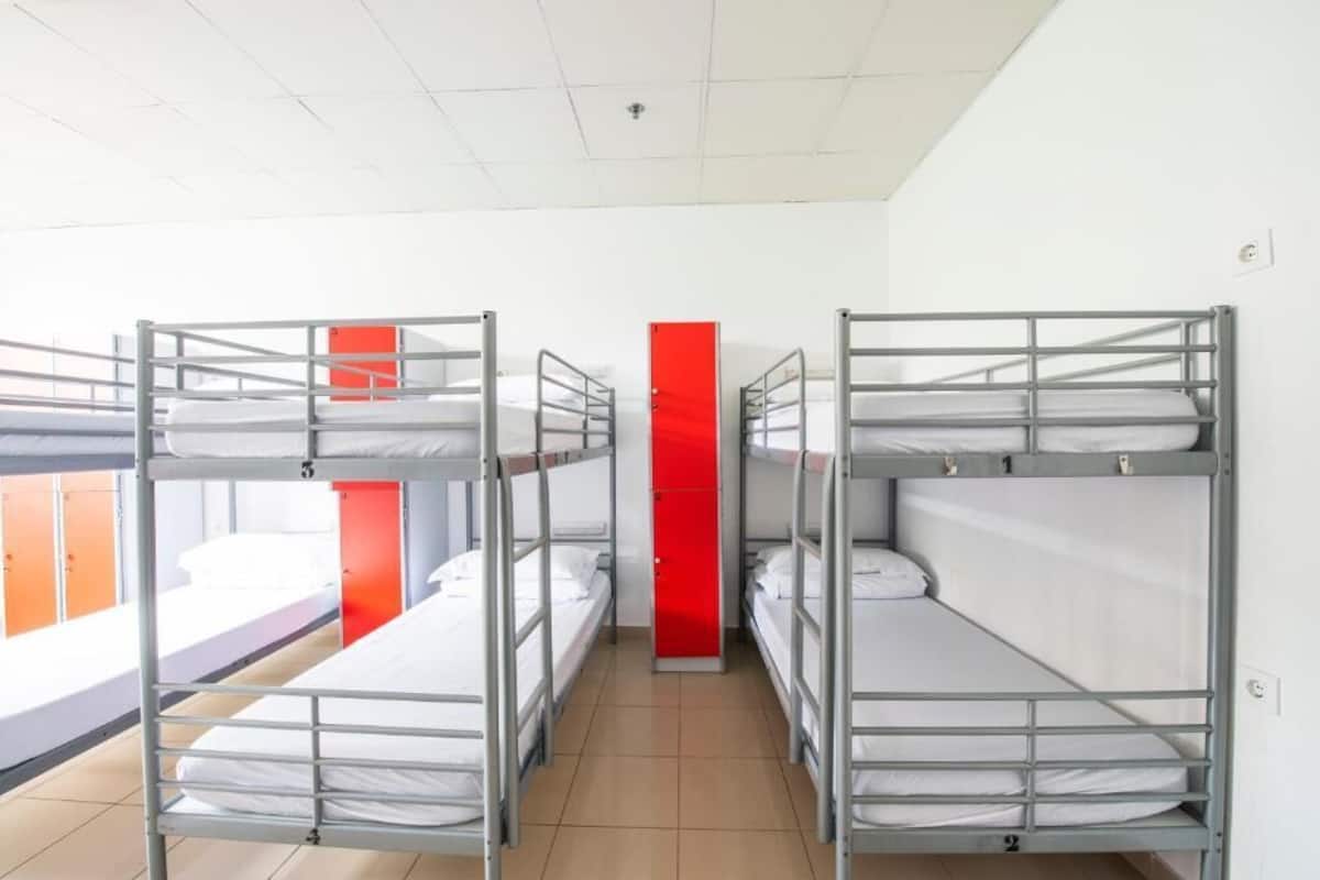 A dormitory room with bunk beds, a red pillar, and a window with orange curtains.