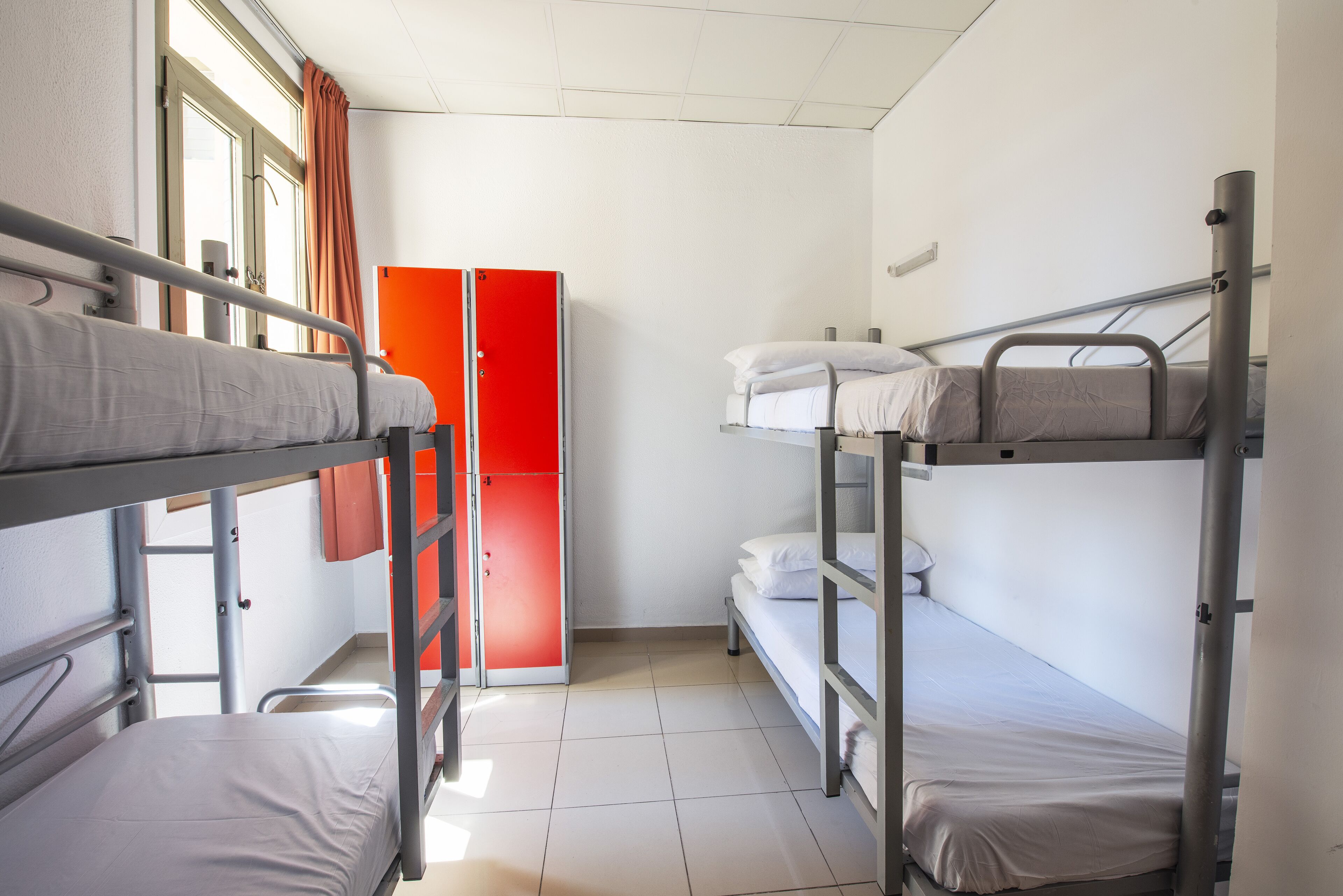 A dormitory room with bunk beds, a red wardrobe, and a window with curtains.