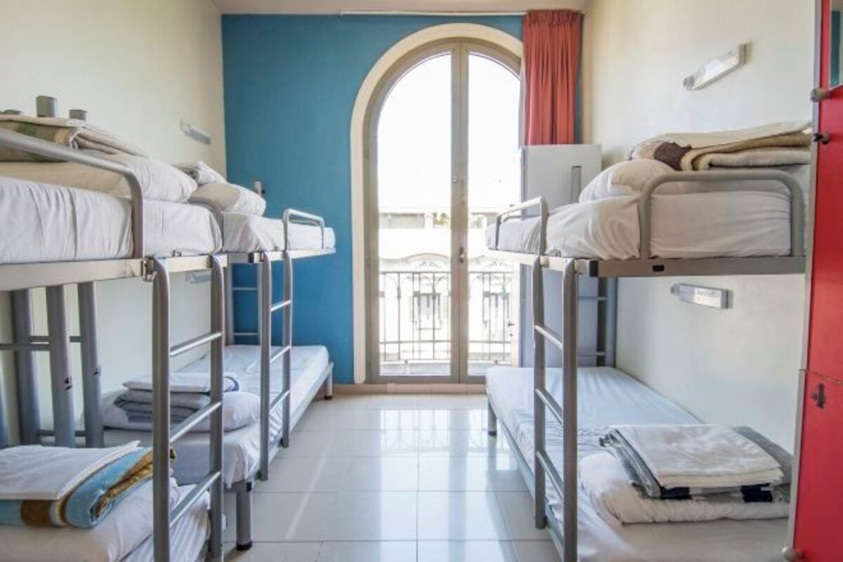 A dormitory room with bunk beds, a door leading to a balcony, and a window with curtains.