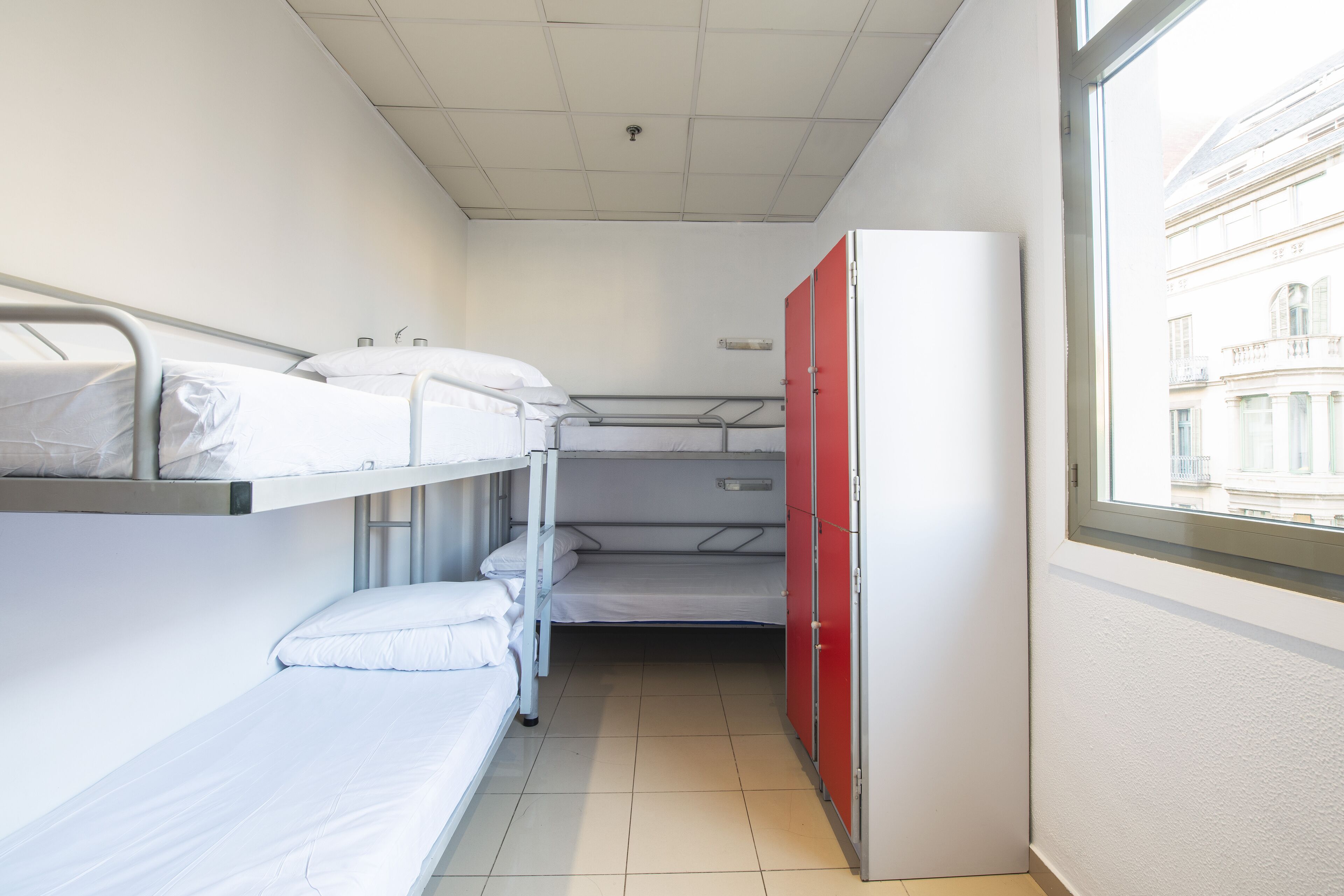 A narrow hotel room with bunk beds, a red wardrobe, and a window with a view of buildings.