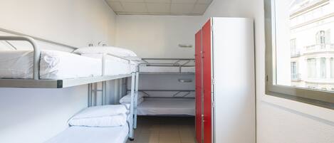 A narrow hotel room with bunk beds, a red wardrobe, and a window with a view of buildings.