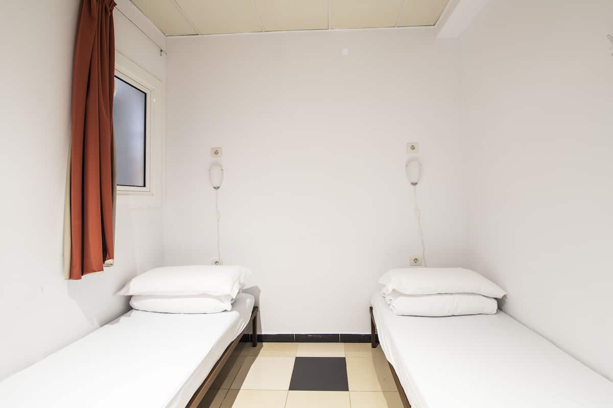 A small, clean room with two single beds, a window with brown curtains, and a tiled floor.