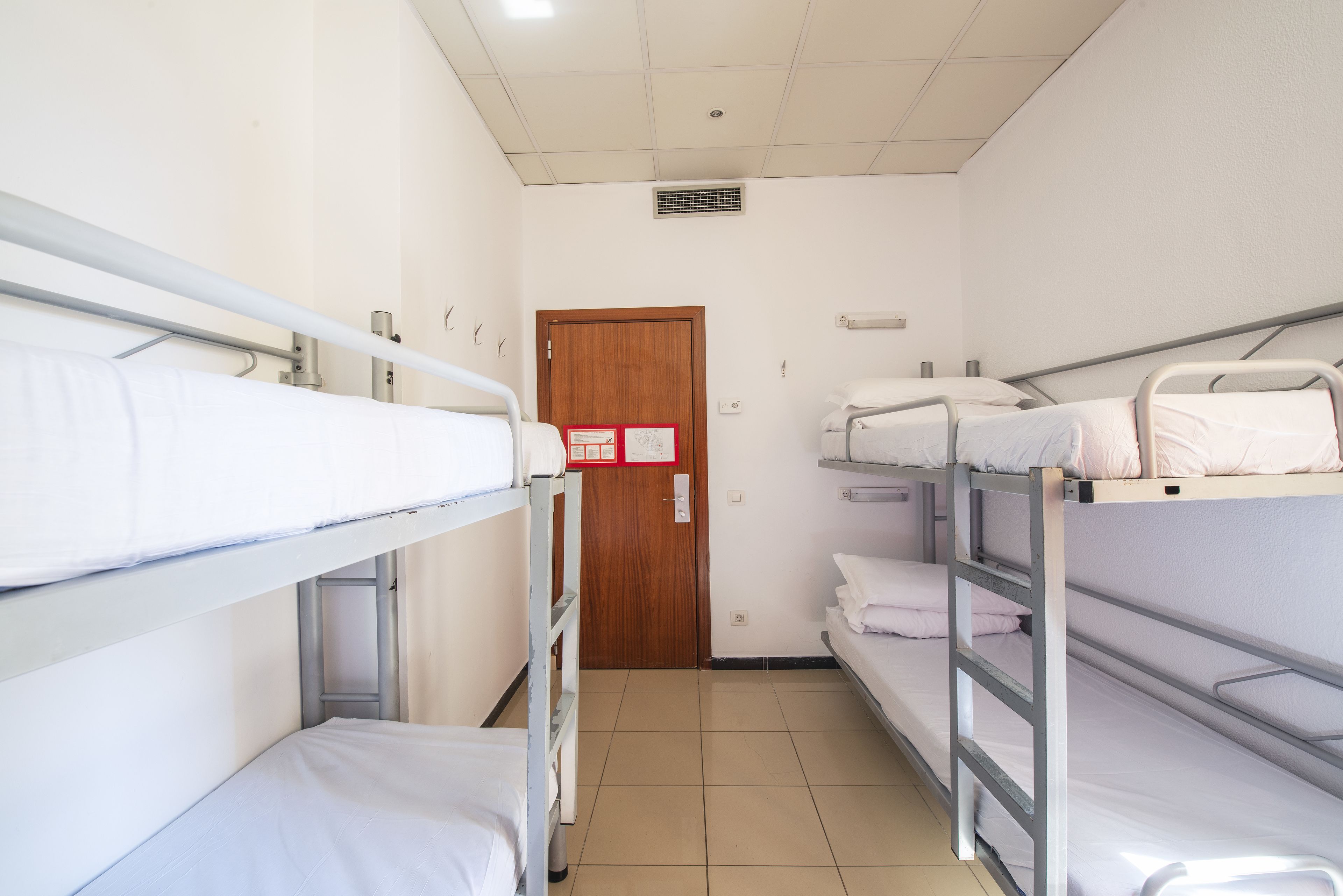 A dormitory room with bunk beds, a wooden door, and a white wall with a red sign.