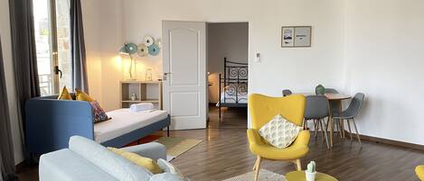 A modern living room with a sofa, a yellow armchair, a dining table with chairs, and a bed with a headboard.