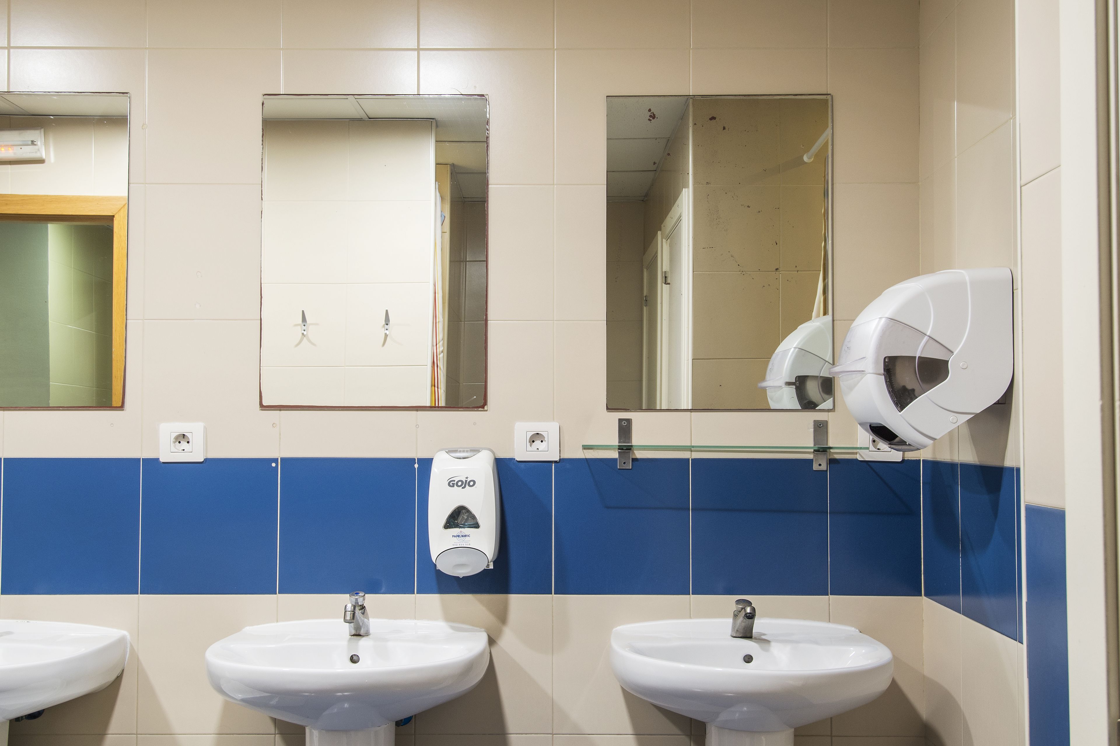 Ein Badezimmer mit drei Waschbecken, einem Handdesinfektionsspender und zwei Spiegeln.