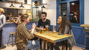 A group of people sitting at a wooden table in a bar with blue accents and hanging pendant lights. - Safestay Barcelona Passeig de Gracia (Barcelona)