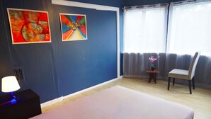 A bedroom with a bed, a chair, a nightstand with a lamp, two paintings on the wall, and a window with curtains. - Stay.plus Budget Paradis (Bergen)