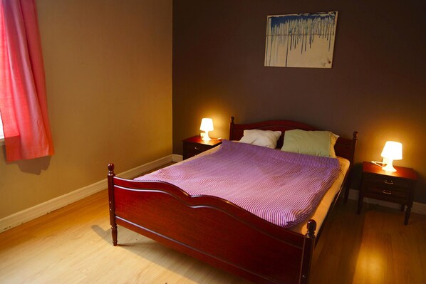 A neatly made bed with a striped blanket, two bedside tables with lamps, a painting on the wall, and a wooden floor. - Stay.plus Budget Paradis (Bergen)