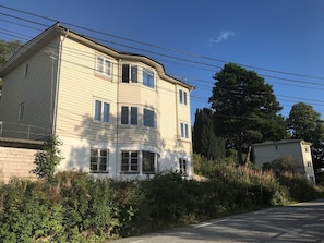 A two-story building with a curved corner, multiple windows, and a visible front yard with dense foliage. - Stay.plus Budget Paradis (Bergen)