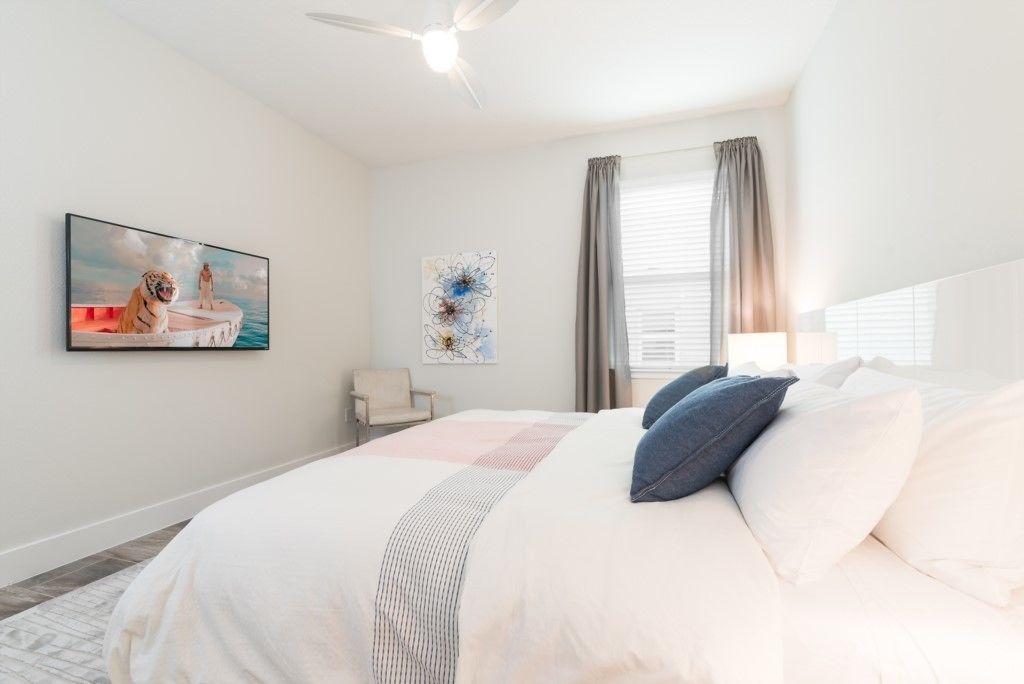 A bedroom with a large bed, a ceiling fan, a window with curtains, and artwork on the walls.