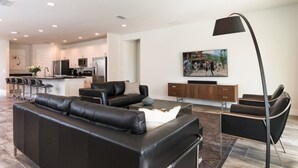 A modern living room with a black leather sofa, a coffee table, and a floor lamp. There is a kitchen in the background with white cabinets and a dining area with bar stools. - Je59003 - The Encore Club Resort - 9 Bed 8.5 Baths Villa (Kissimmee)