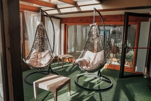 A room with two hanging chairs, a small table, and a view through a glass door. - Hotel La Soldanella (Moena)