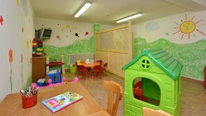 A child's playroom with a green playhouse, a table with chairs, and a bookshelf with toys. - Hotel La Soldanella (Moena)