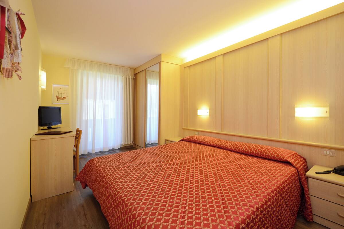 A hotel room with a bed, a desk with a computer, a chair, a wardrobe, and a mirror.
