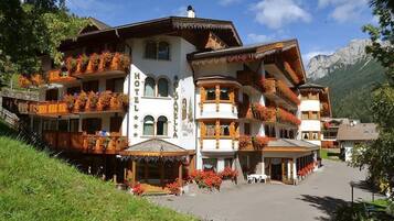 Hotel com várias varandas adornadas com flores, entrada central e paisagem montanhosa ao fundo.