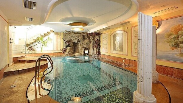 A large indoor swimming pool with a stone wall backdrop, a staircase leading to an upper level, and a ceiling with recessed lighting. - Hotel La Soldanella (Moena)