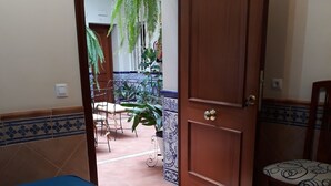 A hotel room with two beds, a wooden door, a chair, and a view of a patio with plants and a tiled floor. - Pensión Doña Trinidad (Seville)