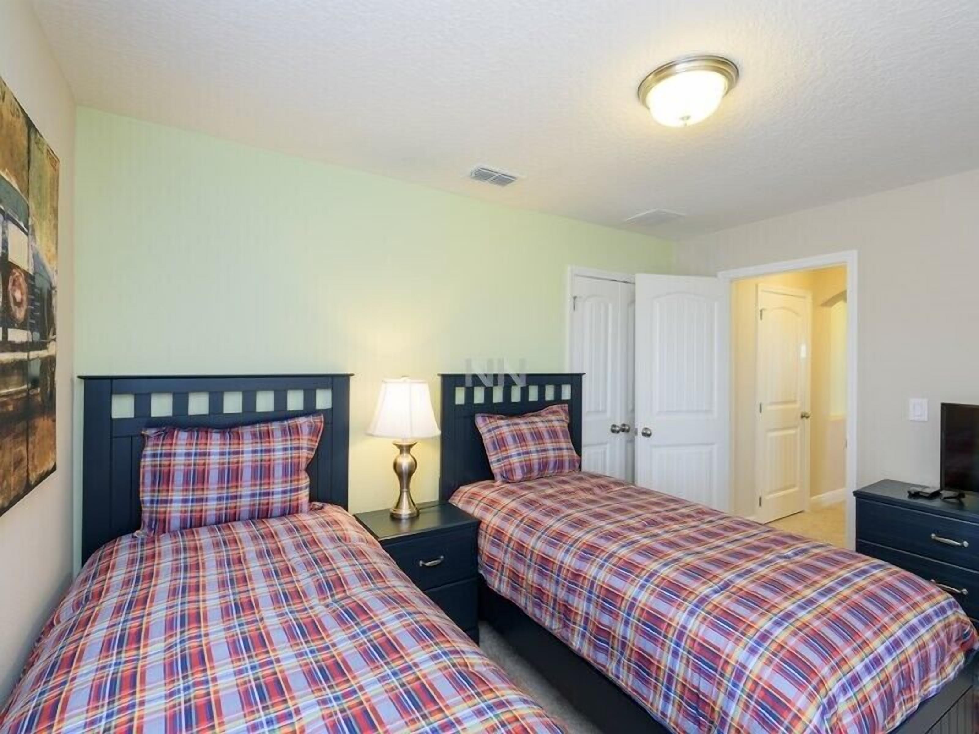 A bedroom with two beds, each with a plaid bedspread, a bedside lamp, and a nightstand. There is a white door and a small TV on a dresser.