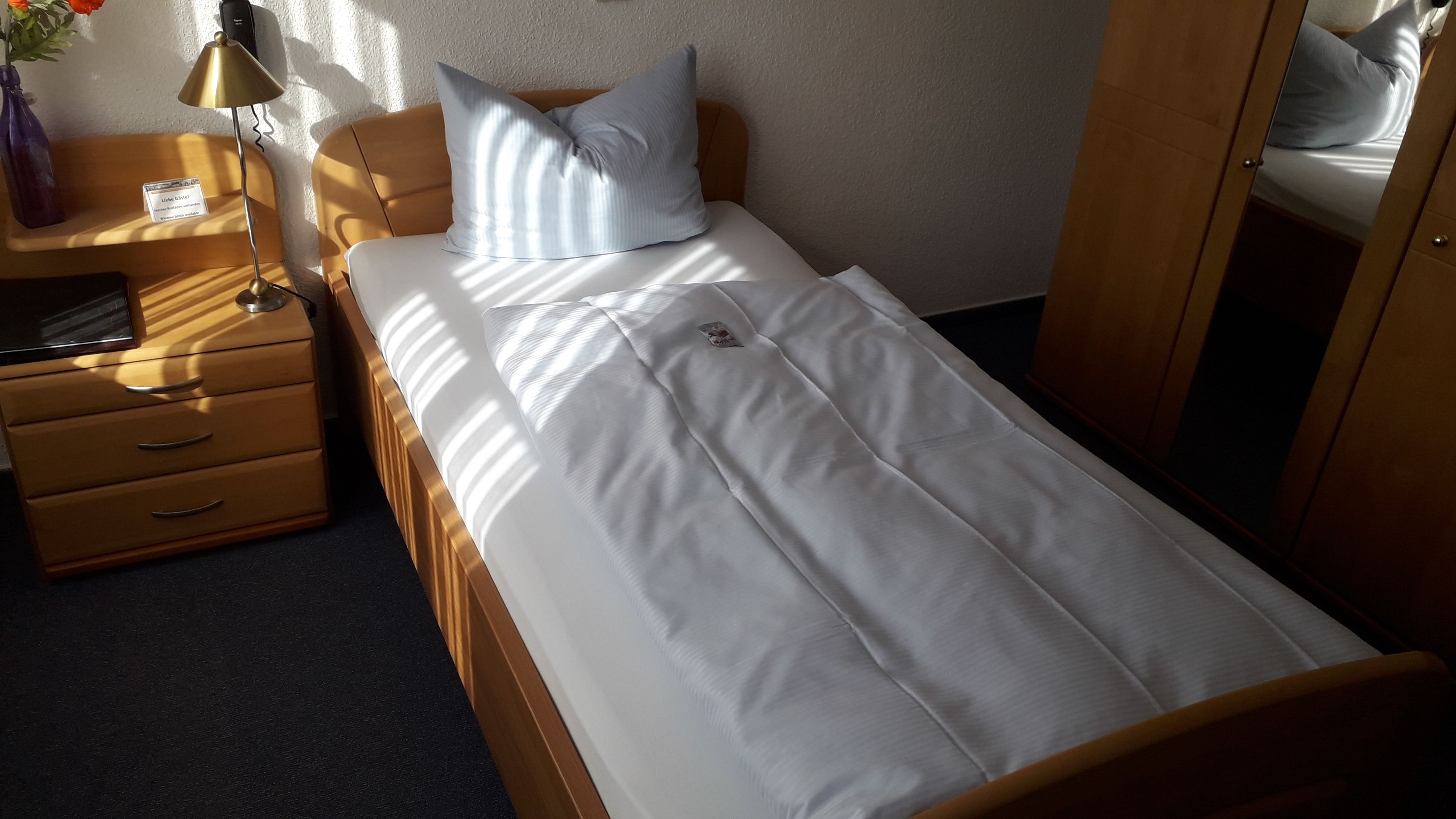 A single bed with white bedding, a wooden headboard, a nightstand with a lamp, and a small table with a vase of flowers.