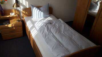 A single bed with white bedding, a wooden headboard, a nightstand with a lamp, and a small table with a vase of flowers.